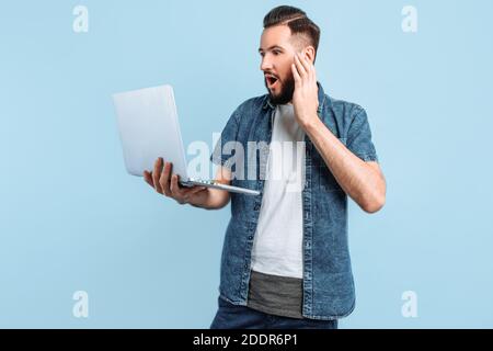 Eine schockierte, überraschte Person hält einen Laptop und schaut überrascht auf den Bildschirm, auf einem isolierten blauen Hintergrund stehend Stockfoto