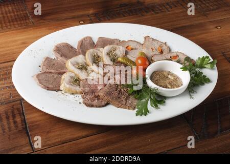 Gebratenes Roastbeef, Hühnerbrötchen, Zunge und Schweinefleisch mit Gemüse auf einem weißen ovalen Teller. Holzhintergrund. Stockfoto