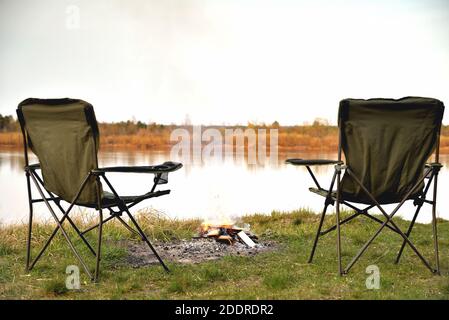Ein Paar Touristensessel, ein Klappstuhl, ein Lagerfeuer am Flussufer am Abend bei Sonnenuntergang, Dämmerung im Herbst bei Sonnenuntergang, Entspannung und Ruhe, Stockfoto