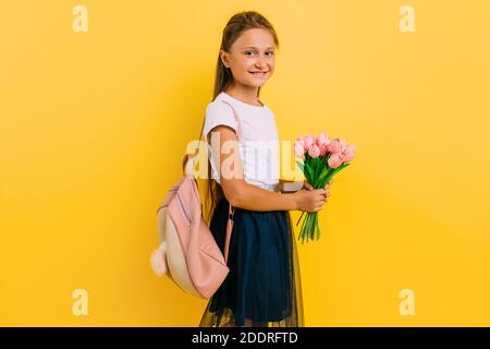 Fröhliches Teenager-Schulmädchen mit einer Aktentasche und einem Blumenstrauß aus Tulpen, die auf einem gelben Hintergrund stehen. Urlaubskonzept Stockfoto