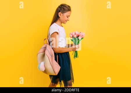 Fröhliches Teenager-Schulmädchen mit einer Aktentasche und einem Blumenstrauß aus Tulpen, die auf einem gelben Hintergrund stehen. Urlaubskonzept Stockfoto