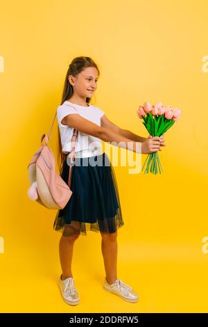 Fröhliches Teenager-Schulmädchen mit einer Aktentasche und einem Blumenstrauß aus Tulpen, die auf einem gelben Hintergrund stehen. Urlaubskonzept Stockfoto