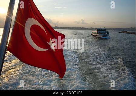 Rote türkische Flagge aus nächster Nähe auf der Fähre gegen Wasser und Wellen im Meer in Bosporus (Bosporus) bei Sonnenuntergang. Schwerpunkt auf anderen Passagierschiffen. Stockfoto