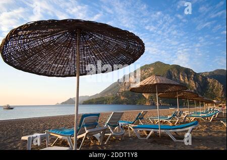 Stroh Strand Sonnenschirme und Liegestühle am Cirali Strand am Morgen bei Sonnenaufgang ohne Menschen. Berge und sonnenbeschienenen blauen Himmel auf dem Hintergrund. Stockfoto