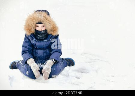 Ein frustriertes Kind mit traurigen Augen in einem blauen Jumpsuit mit einer Pelzhaube in Fäustlingen und einer Gamasche anstelle eines Schals, der ruddige Wangen vor Frost schützt. Stockfoto