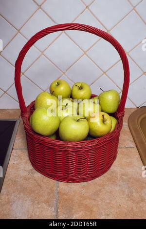 Mehrere grüne Äpfel in einem roten Korbkorb ruhen auf einer rustikalen und weiß gefliesten Küche. Stockfoto