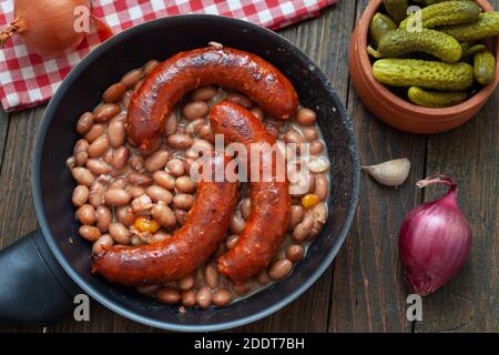 Gebackene Bohnen mit Würstchen und Gemüse in der Pfanne. Tavche na gravche, traditionelles mazedonisches Gericht auf Holztisch. Stockfoto