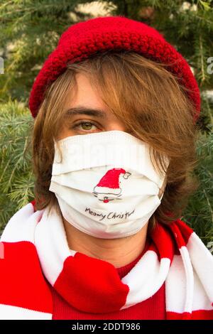 Junger Mann feiert Weihnachten mit schützenden medizinischen Gesichtsmaske wegen covid-19 Pandemie. Junger Mann zieht weihnachtsmann Hut auf eine Gesichtsmaske und feiert C Stockfoto