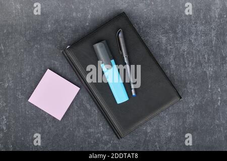 Kugelschreiber und ein Marker liegen auf einem schwarzen Tagebuch, neben Papierstäbchen für rosa Schallplatten, Business Stillleben Stockfoto