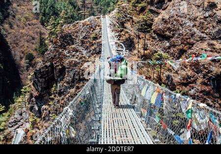 Nepalischer Porter trägt schwere Last auf dem Rücken und geht die Brücke entlang zum Everest-Basislager. Himalaya. Nepal. Stockfoto