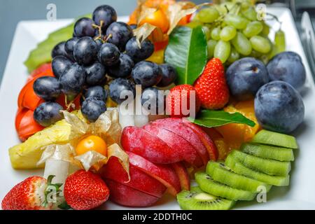 Auf einer Platte stehen verschiedene Obstscheiben, schwarze und weiße Trauben, Kiwi, Pflaumen, Ananas, Erdbeeren und Physalis. Der Blick von oben. Stockfoto
