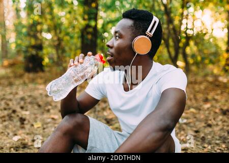 Ein junger, sportlicher Afroamerikaner entspannt sich nach einem intensiven Training. Ein Mann ruht sich aus und trinkt Wasser, während er nach einem Mor auf dem Boden im Wald sitzt Stockfoto