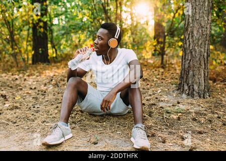 Ein junger, sportlicher Afroamerikaner entspannt sich nach einem intensiven Training. Ein Mann ruht sich aus und trinkt Wasser, während er nach einem Mor auf dem Boden im Wald sitzt Stockfoto