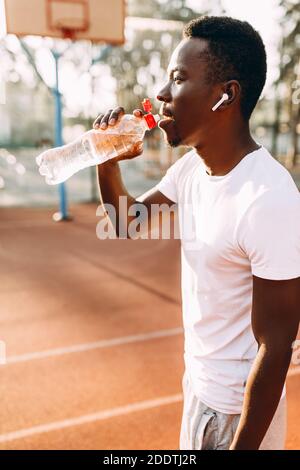 Ein junger, sportlicher Afroamerikaner entspannt sich nach einem intensiven Training. Eine Person hört Musik in Kopfhörern und trinkt Wasser Stockfoto