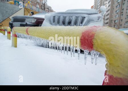 Gelb-roter Parkzaun in einer eisigen Kruste mit Eiszapfen. Vor dem Hintergrund der SUVs. Stockfoto