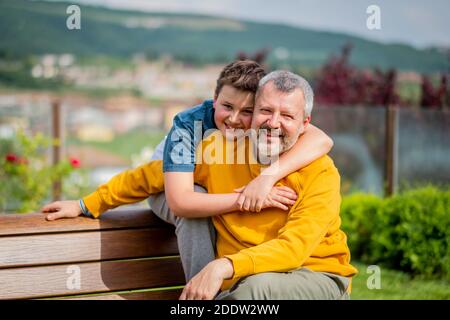 Vater und Sohn umarmen sich und genießen gemeinsam einen sonnigen Tag in der Freizeit auf einer Bank - Vater und Sohn umarmen sich glücklich Stockfoto