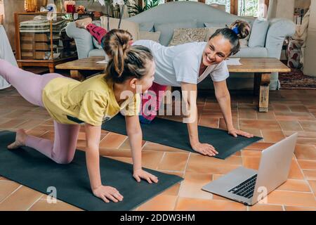 Mutter und Tochter üben online Yoga-Kurse zu Hause während der Quarantäne während der Coronavirus-Pandemie. Familie macht Sport zusammen online Stockfoto