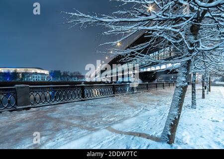 Blick auf die Brücke über den Moskva Fluss durch die Äste von Bäumen in der Nacht im Winter. Die Bäume sind mit Neuschnee bedeckt. Stadtbeleuchtung Stockfoto