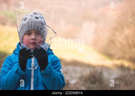 Fünf Jahre alter Junge hält kleines Stück Eis im Wald gegründet. Blick durch den Eisberg zur Kamera. Leerzeichen für Text. Konzept für den kommenden Winter. Stockfoto