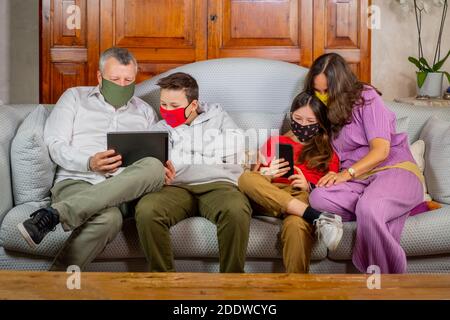 Mutter und Vater beobachten mit ihren Kindern das Tablet und Das Smartphone auf dem Sofa zu Hause - schöne Familie Tragen von Gesichtsmasken gegen Coronavirus Stockfoto