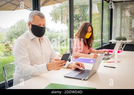 Mutter und Vater arbeiten mit Laptop zu Hause mit Schutzmaske - Mann und Frau arbeiten im Büro Mit Schutzmaske und Sicherheitsdistanc Stockfoto