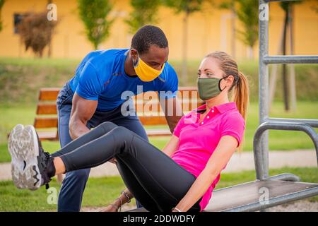 Afro american Fitness-Trainer Coaching helfen Frau tun Liegestütze auf Horizontale Stange mit medizinischem Schutz im Park kreuzen Masken - Youn Stockfoto