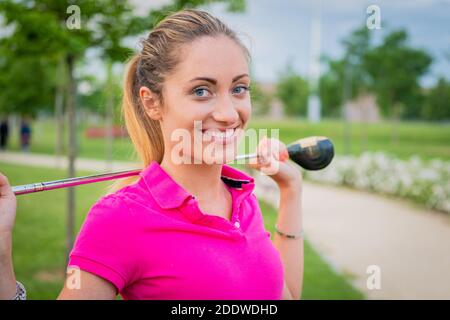 Porträt einer schönen und lächelnden blonden Frau, die ist Golf spielen auf einem Hintergrund von grünen Feld im Freien - Junges Mädchen mit Golfschläger Stick auf dem Rücken Stockfoto