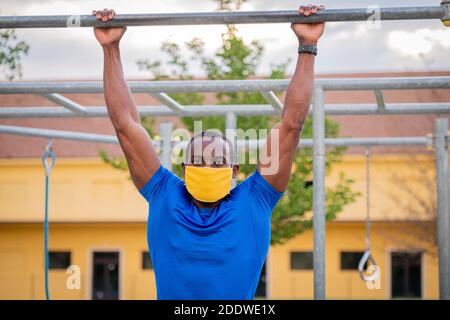 Athlet tut Pull-up auf horizontale Stange mit Schutzmaske - Fitness im Freien im Park während Quarantäne - Afro or afroamerikanischer Mann im Freien pu Stockfoto