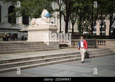 Eine blinde Person, die eine Maske trägt, geht vor eine der Zwillingslöwen-Skulpturen, die vor der New York Public Library in Midtown sitzen. Stockfoto