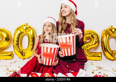 Junge Weihnachtsfamilie, Mama und Tochter in Weihnachtsmützen, hält Kisten Popcorn, sitzen mit Weihnachtsgeschenken, auf einem weißen Hintergrund mit Ballons in Stockfoto