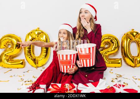 Junge Weihnachtsfamilie, Mutter und Tochter in Weihnachtsmützen halten Kisten mit Popcorn, schockiert beim Anblick eines Films mit Weihnachtsgeschenken sitzen, auf einem weißen Bac Stockfoto