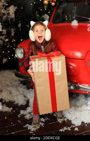 Das Mädchen ist glücklich mit einem großen Neujahrsgeschenk. Kind mit einem Weihnachtsgeschenk in den Händen. Glückliches kleines Mädchen auf dem Hintergrund des Neujahrs Dekor. Stockfoto