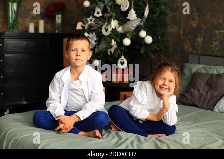 Kinder sitzen auf dem Bett vor dem Weihnachtsbaum. Junge und Mädchen warten auf weihnachten Stockfoto