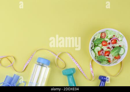 Gesunde ausgewogene Ernährung Konzept, frischer Salat mit Gurken Tomate, Maßband, Hantel, Flasche Wasser auf gelbem Hintergrund. Draufsicht copy space Stockfoto
