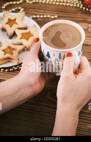 Weibliche Hand halten Tasse heiße Schokolade oder Schokolade mit Auf Holztisch mit Lebkuchenkuchen und Weihnachtsdekoration Stockfoto