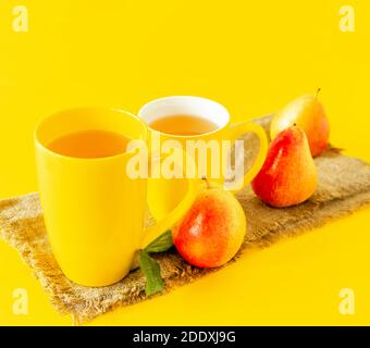 Zwei Tassen Birnensaft und reife Birnen auf Quappen-Tuch und gelbem Hintergrund. Stockfoto