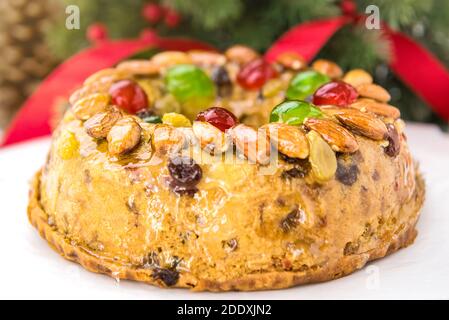 Nahaufnahme von glasierten runden hohlen bunten Weihnachtsfruitcake gekrönt Mit Mandeln und Glace Kirschen auf weißem Teller mit Grün Kiefernnadeln und rote Rippe Stockfoto