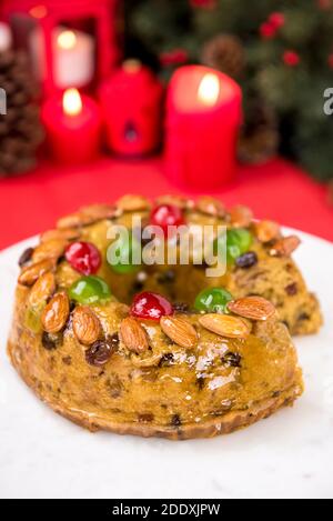 Glasierte bunte Weihnachten Obstkuchen gekrönt mit Mandeln und Glace Kirschen Auf weißem Teller mit Kerzen und Dekorationselementen im Hintergrund Stockfoto