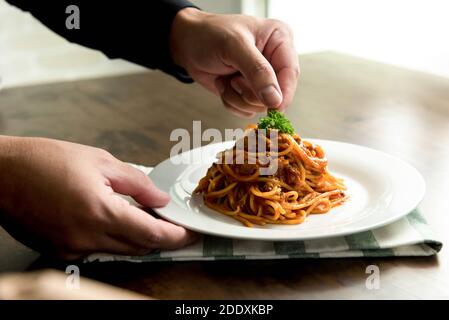 Koch setzt Petersilie auf köstliche Spaghetti Bolognese in Ein weißer Teller auf dem Küchentisch Stockfoto