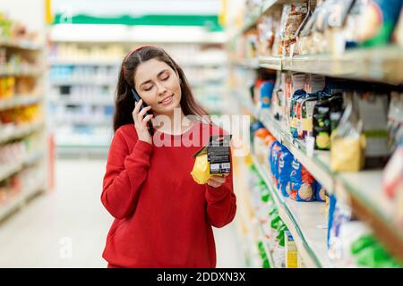 Eine junge hübsche Frau hält eine Packung Getreide und ruft auf ihrem Smartphone an. Supermarkt Halle im Hintergrund. Das Konzept der mobilen Kommunikation Stockfoto