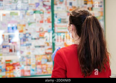 Eine Brünette in einem roten Pullover wählt Medikamente in einer Apotheke.im Hintergrund ist das Fenster verschwommen. Rückansicht. Das Konzept des Kaufens von Medicin Stockfoto