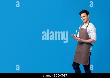 Lächelnder junger, hübscher Barista mit Schürze und Schreibbefehl auf der Zwischenablage Isoliert auf blauem Hintergrund Stockfoto