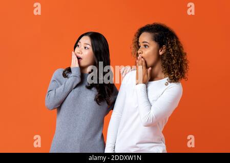 Asiatische und afroamerikanische Frau Freunde in schockiert Geste mit Hände bedecken Mund isoliert in orange Hintergrund Stockfoto