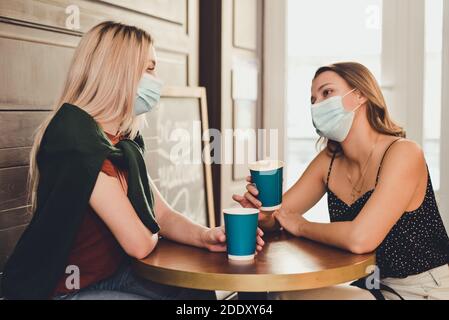 Zwei schöne Mädchen mit Masken auf ihren Gesichtern verbringen Zeit zusammen in Kaffee und Tee trinken. Schutz vor Coronavirus Covid-19 während der pande Stockfoto