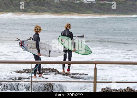 Zwei junge männliche Surfbrettfahrer beobachten sorgfältig, wie sie sich vorbereiten Um in das Wasser von den Felsen bei Schwarz zu springen Head Beach Ocean Pool in Australien Stockfoto