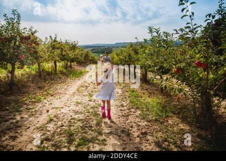 Das Mädchen im Hut läuft mit süßem Apfel zu ihrem Vater und der kleinen Schwester im Apfelgarten. Herbst. Sommer. Ungarn Stockfoto