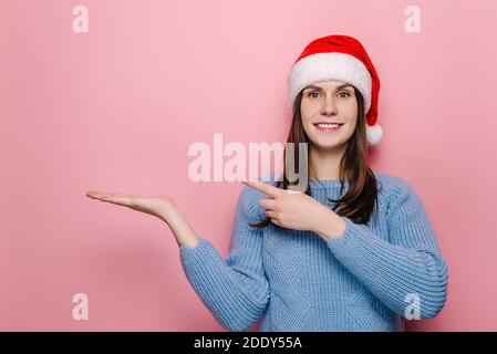 Attraktive junge Mädchen hält Palm angehoben, Punkte mit Zeigefinger gegen rosa Wand für Werbung, in Weihnachten roten Hut gekleidet Stockfoto