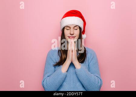 Zufrieden gut aussehende Frau in Weihnachten roten Hut hält Palmen in betende Geste, hat positiven Ausdruck, sanftes Lächeln, macht Wunsch Stockfoto