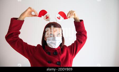 Kaukasische Frau in einem roten Mantel und einer medizinischen Maske wird mit Santa Claus Stirnband gespielt. Stockfoto