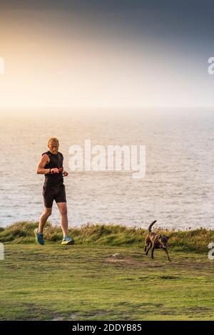 Ein Mann, der seine Zeit beim Joggen auf einem Fußweg mit seinem Hund am Pentire Point East in Newquay in Cornwall überprüft. Stockfoto